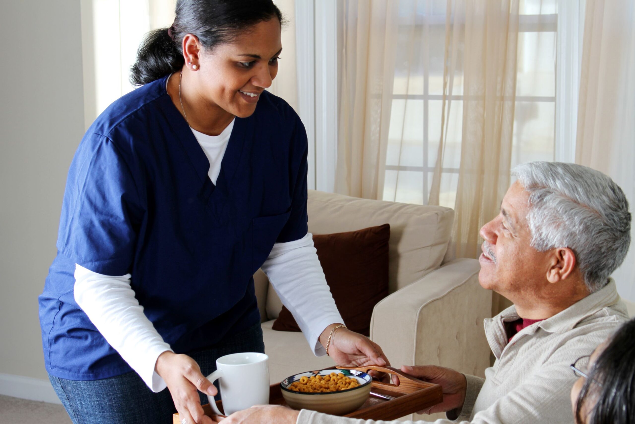 Healthcare worker in blue and white scrubs places a food tray with cereal and a white coffee mug in front of an older gentleman with silver hair wearing and ivory colored shirt.