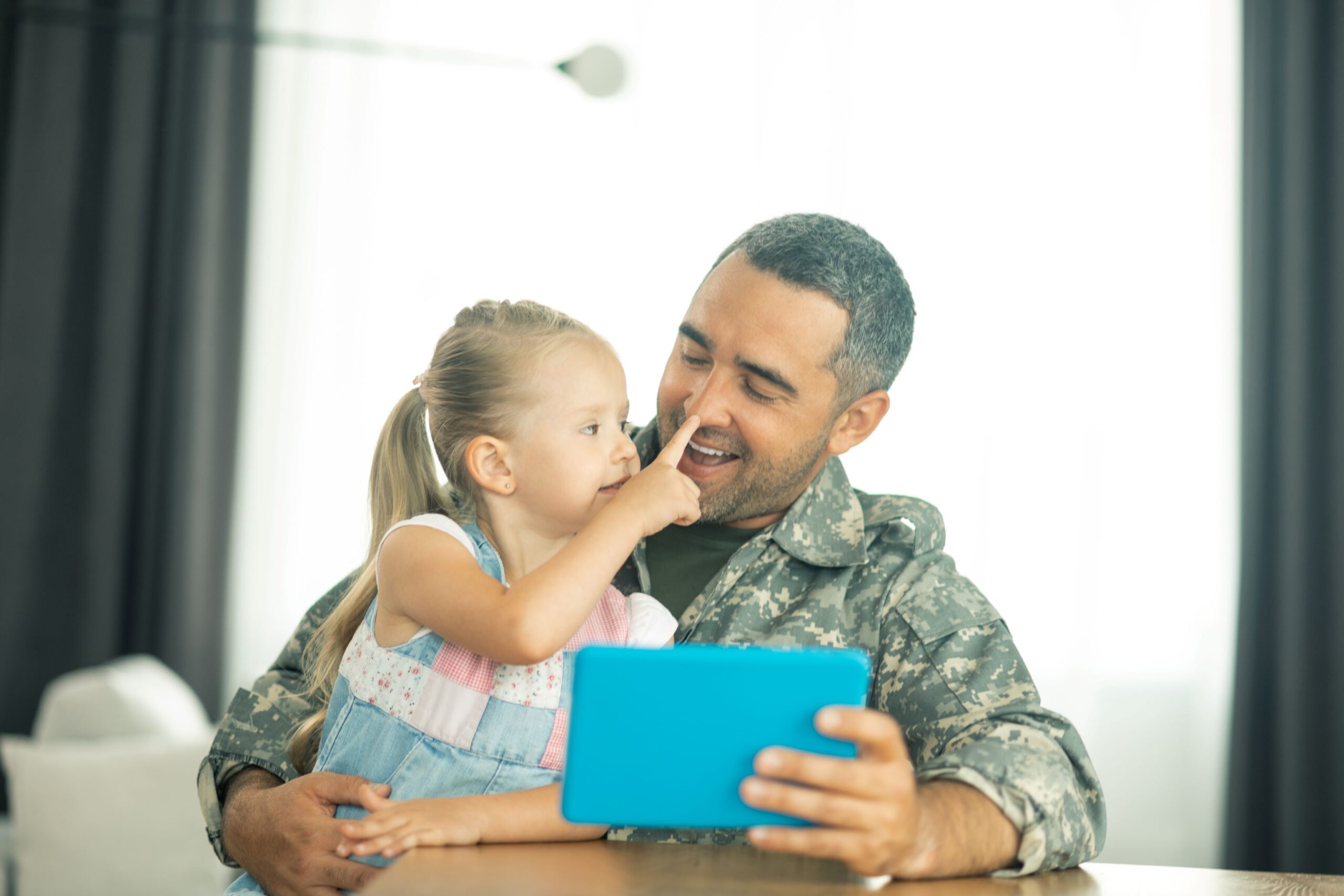 A military dad sits a table with his daughter on his lap. She is touching the tip of his nose with her finger and while he is holding a blue iPad.